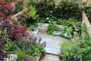 A landscaped garden with stone paving, brick walls, various green plants, ferns, and flowering perennials among the shrubs, featuring a small seating area and potted plants.