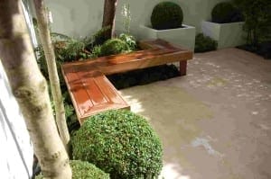 A wooden L-shaped bench is positioned in a bespoke garden with neatly trimmed round bushes and square planters on a light-colored paved surface.