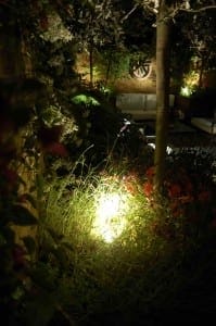 A garden at night with lush greenery, red flowers, and a bright ground light illuminating the plants. Outdoor lighting highlights the scene, while a cozy seating area is visible in the background.