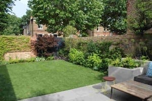 A small backyard garden with fake lawns, various green plants, a brick wall, a wooden bench with cushions, a small round table, and paved patio tiles.