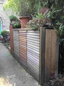 A wooden binshed with horizontal slats stands outdoors, topped with potted plants and surrounded by lush greenery.