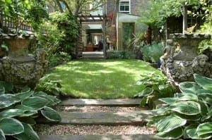 A well-maintained garden with a grassy lawn, stone steps, large ornate planters, vibrant green plants including a Himalayan birch, and a house in the background.