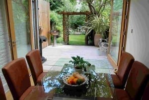 Dining table with brown chairs and a fruit bowl is set near open glass doors, leading to a garden with greenery, Himalayan birch trees, and a wooden pergola outside.
