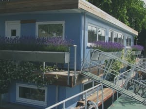 Blue houseboat with large windows and flower boxes of lavender and white flowers overlooks oval gardens; bicycles are parked alongside the dock.