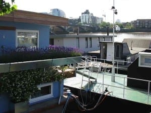 Several stylish houseboats are moored along a river, with buildings and a bridge visible in the background on a clear day.