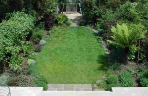 A neatly mowed rectangular lawn is bordered by various shrubs, flowers, and trees, with hints of oval gardens and a small play area alongside charming garden structures in the background.