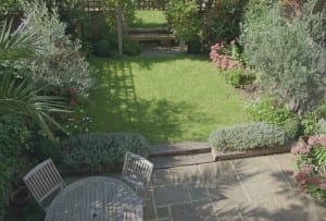 View of a small, neatly maintained oval garden with a lawn, surrounding flower beds, a wooden bench at the back, and a stone patio with a round table and chairs in the foreground.