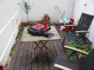 A small wooden patio with a folding table holding a backpack and camera, surrounded by black chairs, potted plants including acers, and cleaning tools against a white wall.