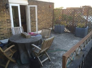 A rooftop patio offers a framed view with a round table draped in cloth, four wooden chairs, large planters, a garden hose, and an open door leading into a brick building.