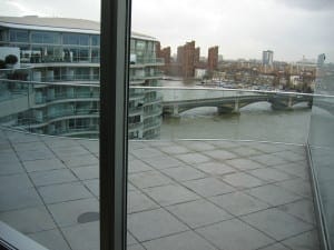 View from a modern glass balcony with a roof garden overlooking a river, featuring an arched bridge and city buildings in the background under a cloudy sky.
