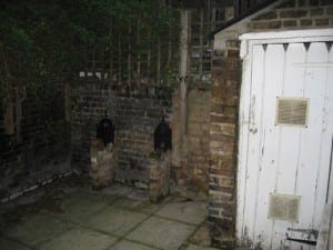 A small outdoor area with stone paving, brick walls, a white wooden door on the right, and two black metal covers on brick stands—an inviting space enhanced by subtle touches of Trompe l’Oeill artistry.