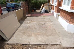 A small front garden with paving stones and exposed bricks, where construction materials and debris are piled near the entrance of a red brick house.