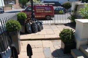 Several black garbage bags and stacked wood are placed on a paved front yard, with two potted shrubs adding a touch of Italianate Garden style and a red van parked on the street in the background.