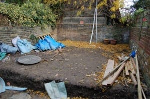 A backyard with bare soil, scattered wooden planks, tarps, plant pots, and fallen leaves. Brick walls and some overgrown plants border the area, with space for a stylish floating bench to enhance the outdoor setting.