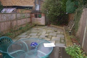 A small paved backyard designed as an outdoor room, featuring a wooden shed, a fence, a round glass table with chairs, and a notebook with a pen on the table. Lush plants and shrubs line the edges for a cozy retreat.
