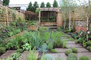 A landscaped garden with stone pathways, various green plants and flowers, wooden fences, recent renovations, and a pergola at the back.