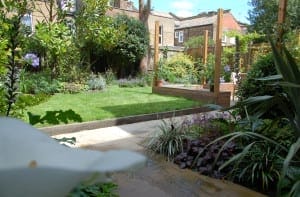 A landscaped Acton backyard garden with a lush lawn, flower beds, wooden posts, and a raised wooden deck, all surrounded by various plants and shrubs.