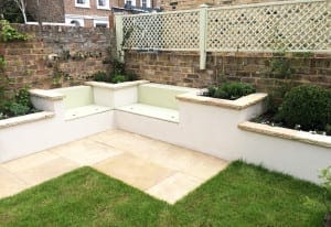 A small garden with a stone patio, grass lawn, built-in corner bench, raised beds as planters, brick walls, and a lattice privacy screen.