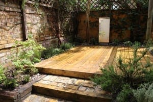 A small courtyard garden features a raised wooden deck, brick paving, climbing plants on the walls, and surrounding greenery for a truly garden rejuvenating atmosphere.