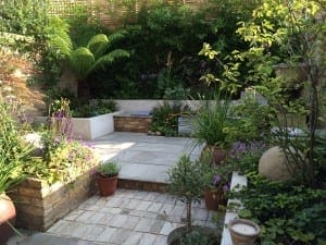 A small, paved garden patio with built-in white benches, exotic potted plants, lush greenery, and a stone sphere decoration.