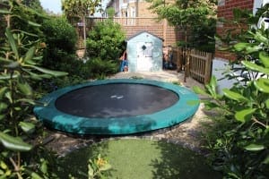 A friendly sunken trampoline with a green edge is set in a landscaped garden surrounded by plants, a small shed, and lush trees and bushes.