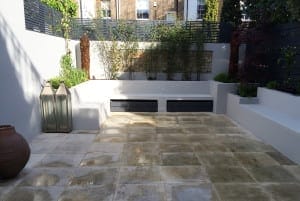 Modern patio with tiled flooring, built-in white seating, tall plants along the back wall—perfect for composting scraps—two large lanterns, and a brown earthen pot on the left side.