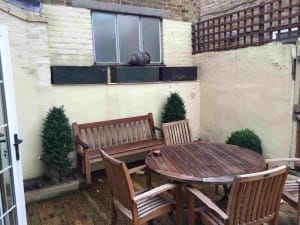 Small outdoor patio with wooden table, four chairs, and a bench; three potted bushes and a watering can on a windowsill against a cream wall in this new rendering.