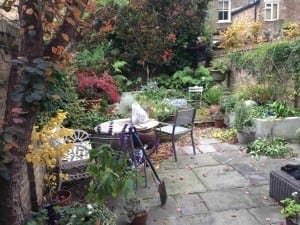 A small garden patio with metal chairs, a table, potted plants, and colorful autumn foliage scattered on the stone floor—perfect for enjoying the season or tackling some autumn maintenance tasks.
