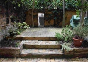 A small, garden rejuvenating space with stone steps leading to a wooden deck, potted plants, lush greenery, and a covered item on the right. A rectangular mirror stands against the back wall.