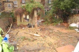 A messy Hammersmith garden area with scattered debris, dirt, bricks, and overgrown plants; walls and trees are visible in the background.