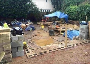 Construction site for a big project with stacked concrete blocks, scattered tools and materials, wooden framework, and a blue tarp shelter over part of the work area, all surrounded by trees and a white building.