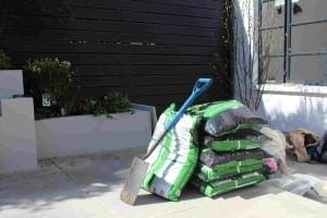 A stack of garden soil bags with a shovel resting on top, placed on a tiled patio near planters and a wooden fence—a perfect setup for composting and nurturing healthy plants.