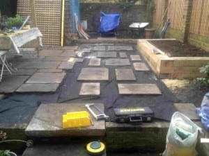 Garden patio under construction in Brook Green, with stone slabs being arranged on a black membrane, surrounded by tools, a raised bed, and garden equipment.