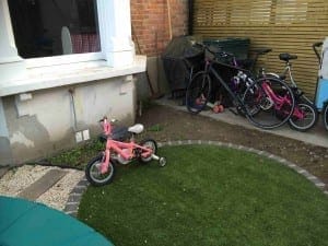 A friendly pink child's bike with training wheels sits on artificial grass beside several larger bicycles near a house and a wooden fence.