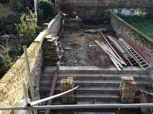 Partially constructed brick patio area with steps, loose bricks, stacked materials, metal pipes, and ladders—signs of ongoing renovations—surrounded by brick walls and some greenery in the background.
