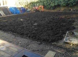 A garden area with freshly turned soil hints at a big project underway, with a small excavator in the background, construction materials on the left, and greenery along the back fence.