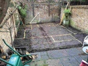 An exotic garden area with marked lines on the soil, surrounded by brick walls, garden tools, and a green wheelbarrow in the foreground.