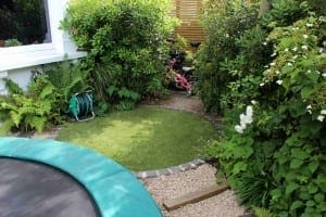 A friendly small garden area with dense green plants, a hose reel, a circular patch of artificial grass, a pink tricycle, and part of a trampoline in the foreground.