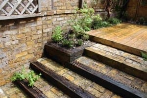 Brick and timber garden steps lead up to a small wooden deck, surrounded by rejuvenating plants growing in beds beside the stairs and along the brick wall.