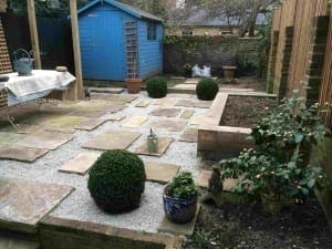 A tidy Brook Green garden with stone paths, gravel, potted plants, trimmed shrubs, a blue shed, and brick walls surrounding the area.