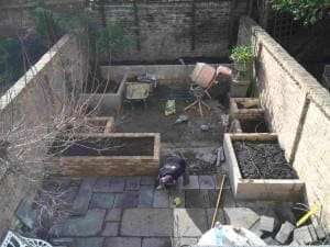 A person is laying exotic stone tiles in a walled garden with raised beds, construction materials, and gardening tools scattered around.