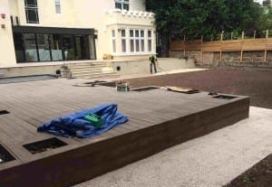 A person tackles a big project in the backyard, working on a newly built wooden deck amid scattered tools and unfinished landscaping near a large house.