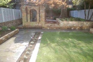 A landscaped Hammersmith backyard features a grass lawn, stone pathway, raised brick planters, and a brick archway with a tree in the background.