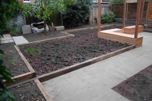 A charming Acton backyard with raised garden beds filled with soil, a paved walkway, and a wooden pergola structure.