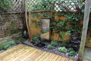 A small, garden-rejuvenating oasis with a wooden deck, lush green plants along the edge, and a tall rectangular water feature set against a brick wall with a wooden trellis.