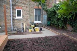 A garden in Acton with tilled soil, tools, yellow bags, and a wheelbarrow sits beside a brick house, where ivy climbs the wall beneath a bright window.