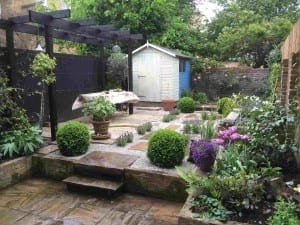 A landscaped garden in Brook Green features stone steps, a pergola, potted plants, a covered table, flowering bushes, and a painted garden shed in the background.