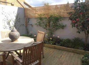 A small garden patio with new rendering on a white wall, wooden decking, a round table and chairs, a decorative lantern, raised flower beds, and climbing plants supported by a wooden trellis.