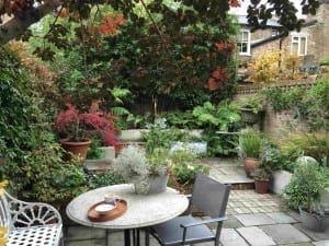 A small, lush garden with various potted plants, a round table with a coffee cup and saucer, grey chairs, and a white bench on a stone patio beside a brick wall—ideal for autumn maintenance or quiet relaxation.