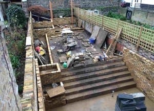 A partially constructed brick patio undergoing renovations features wide steps, building materials, tools, and piles of bricks scattered around, all surrounded by brick walls and wooden trellis fencing.